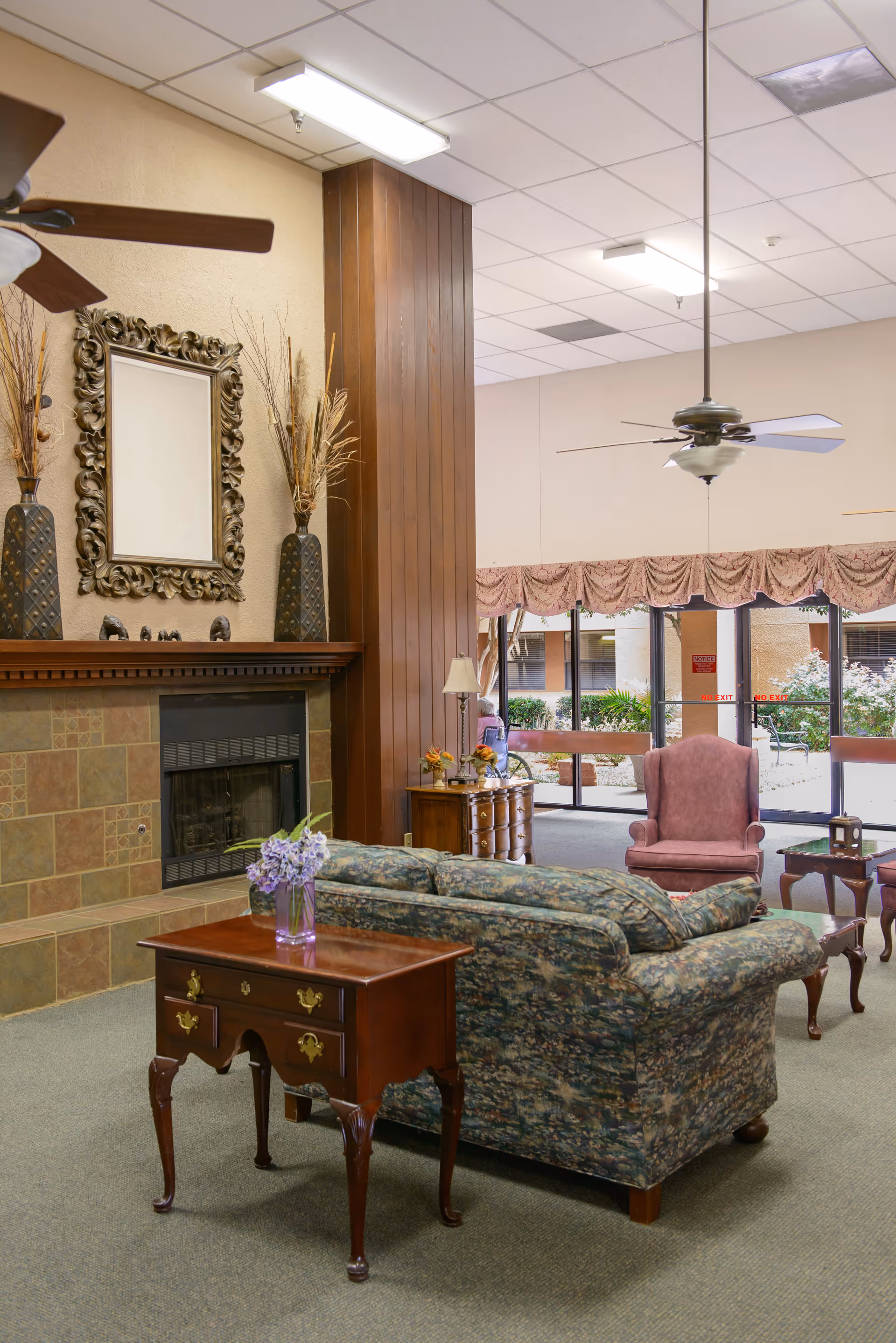 A cozy living room area in a senior care facility featuring a floral patterned sofa, a wooden side table with a vase of purple flowers, a fireplace with a decorative mirror above it, two large vases with dried plants, a pink armchair, ceiling fans, and large windows with draped curtains overlooking an outdoor garden area.