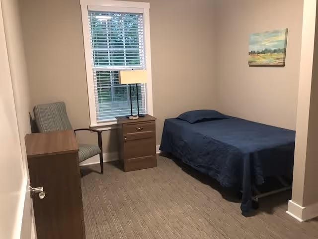 A small, simple bedroom with a single bed covered in a dark blue bedspread, a wooden nightstand with a lamp, a wooden dresser, a striped armchair, and a window with white blinds. A landscape painting hangs on the wall.