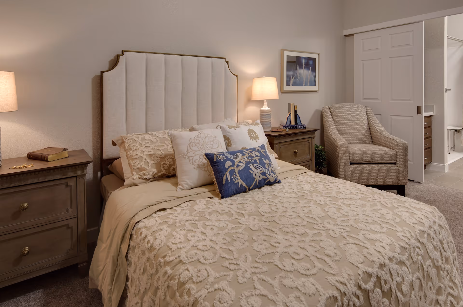 A neatly made bedroom with an upholstered headboard, bedside tables and lamps, decorative pillows, an armchair, and sliding closet doors.