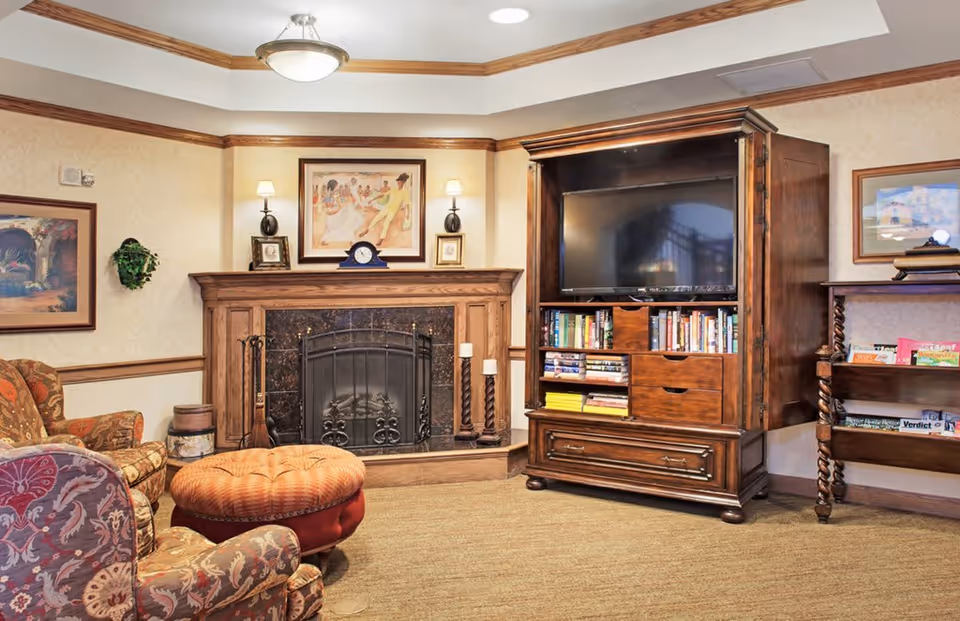 Cozy living room area with a wooden fireplace mantel, a TV inside a wooden cabinet, bookshelves with books and games, patterned armchairs, and a round tufted ottoman. The walls are decorated with framed artwork and sconces with lamps.