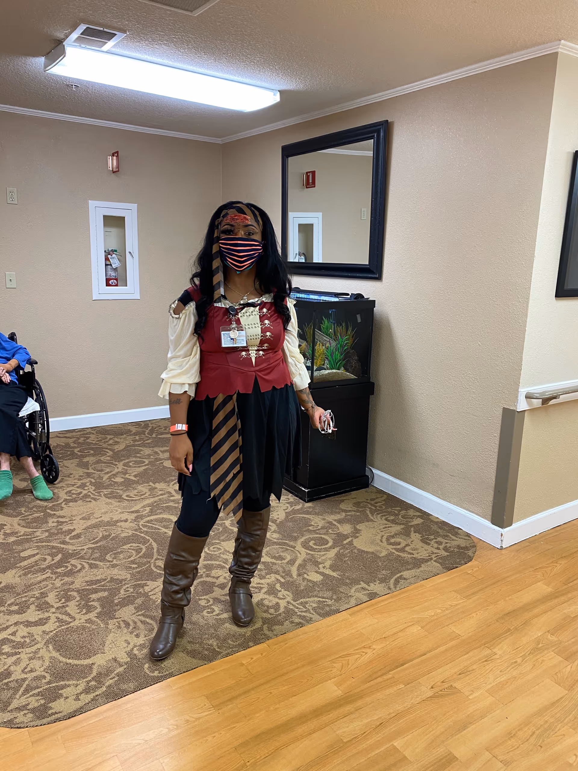 A person dressed in a pirate costume with a striped face mask stands in a hallway of a senior living facility. The hallway has beige walls, a patterned carpet, and a wooden floor. There is a large mirror on the wall behind the person and a fish tank beneath the mirror. Another person in a wheelchair is partially visible in the background.