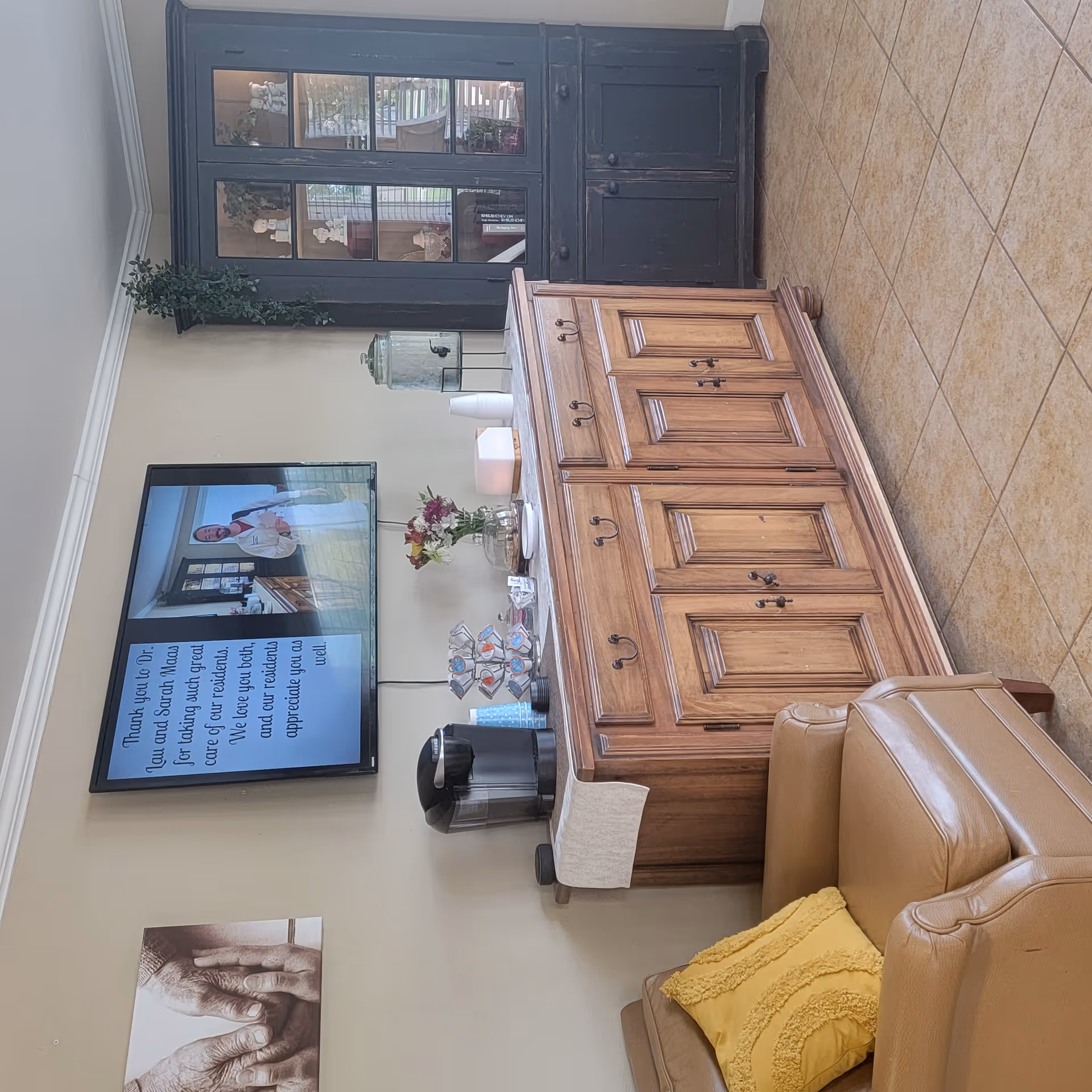 Reception lounge with a wall-mounted TV, a wooden sideboard holding a coffee station and flowers, a glass-front cabinet, and tan leather seating with a yellow pillow.