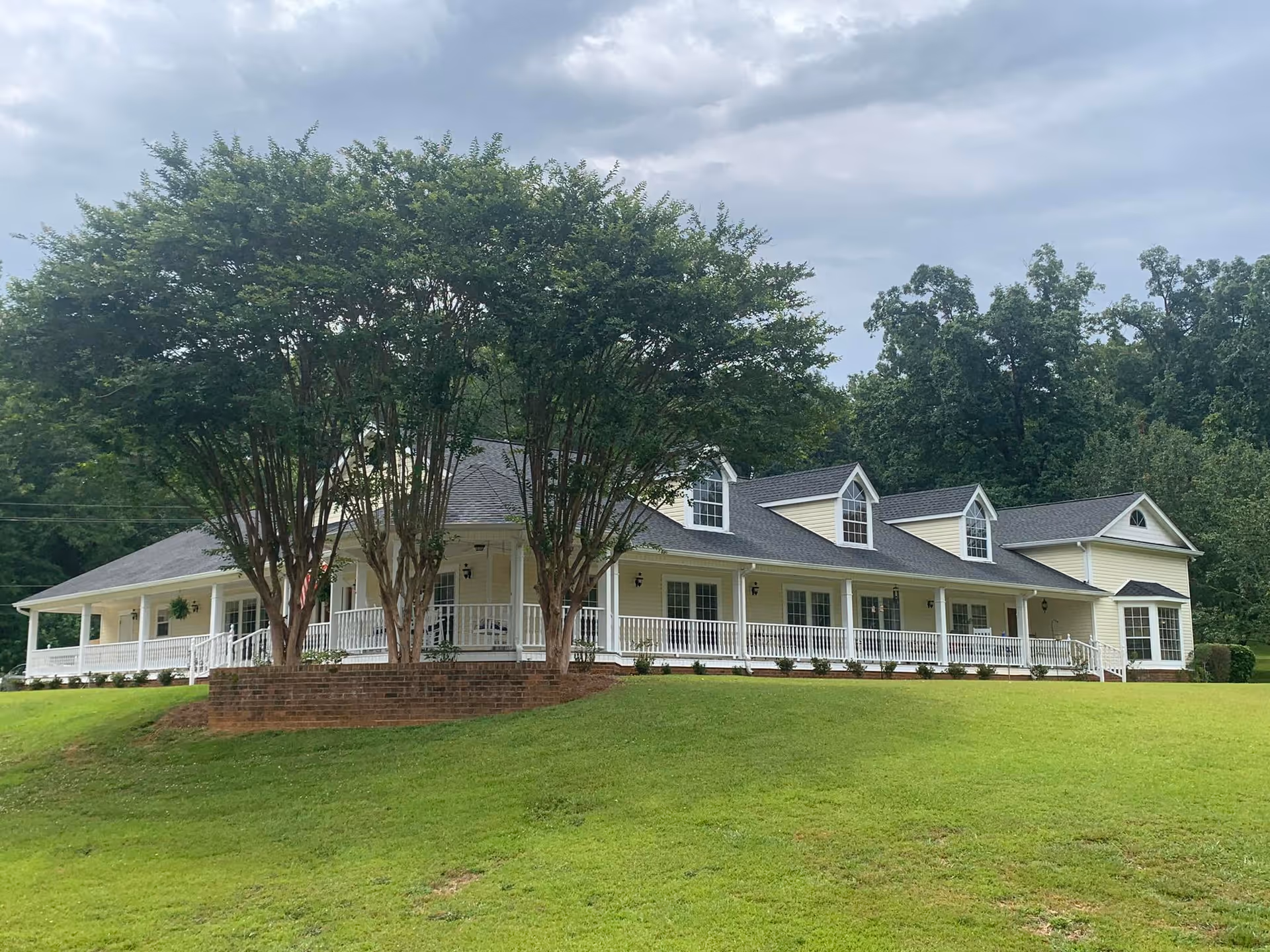 Yellow assisted living building with a long white wraparound porch, dormer windows, and trees on a grassy hill under a cloudy sky.