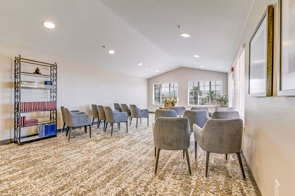 A bright room with a patterned carpet and several gray upholstered chairs arranged in rows facing a window. There is a metal bookshelf on the left side holding books and decorative items. The room has white walls, recessed ceiling lights, and two framed pictures on the right wall.