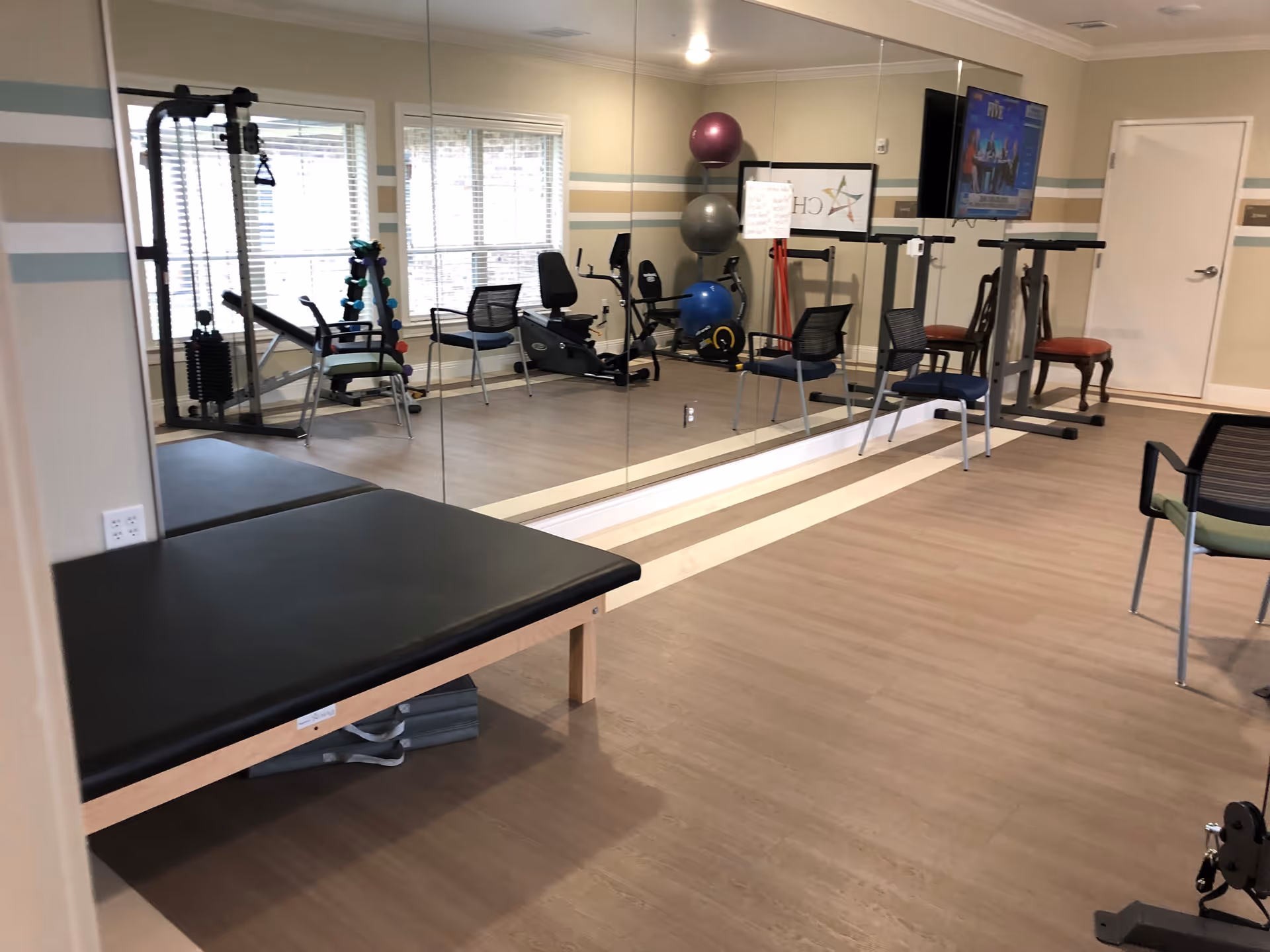 A fitness room with exercise equipment including a weight machine, stationary bike, exercise balls, and dumbbells. There are several chairs arranged around the room, a large mirror on one wall, and a television mounted on the wall. The room has light-colored walls with horizontal stripes and wood flooring.