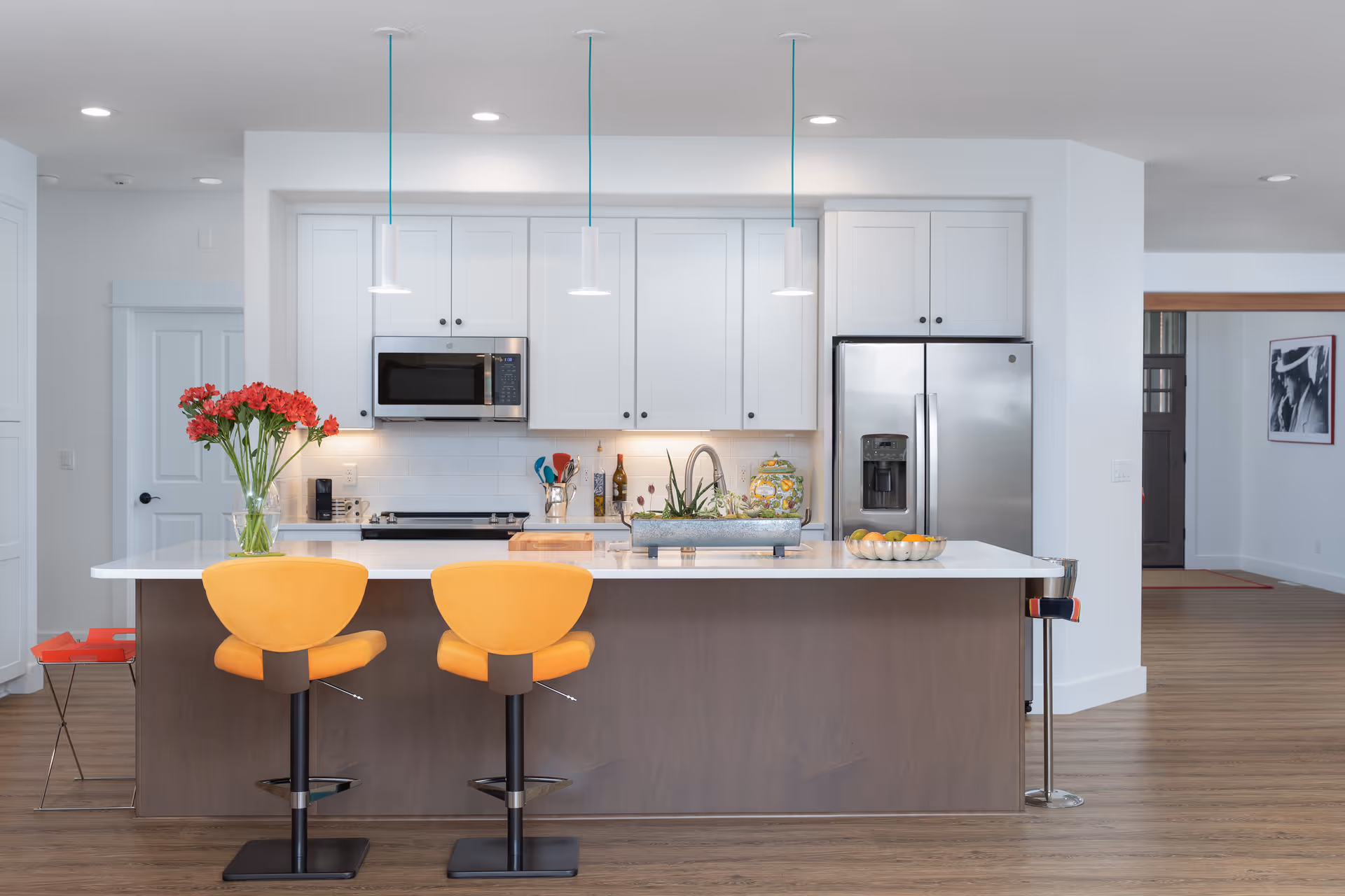 Modern kitchen with white cabinets, stainless steel refrigerator and microwave, a white countertop island with two yellow bar stools, a vase with red flowers, and decorative items on the counter. The kitchen is well-lit with three pendant lights hanging above the island and has a wooden floor.