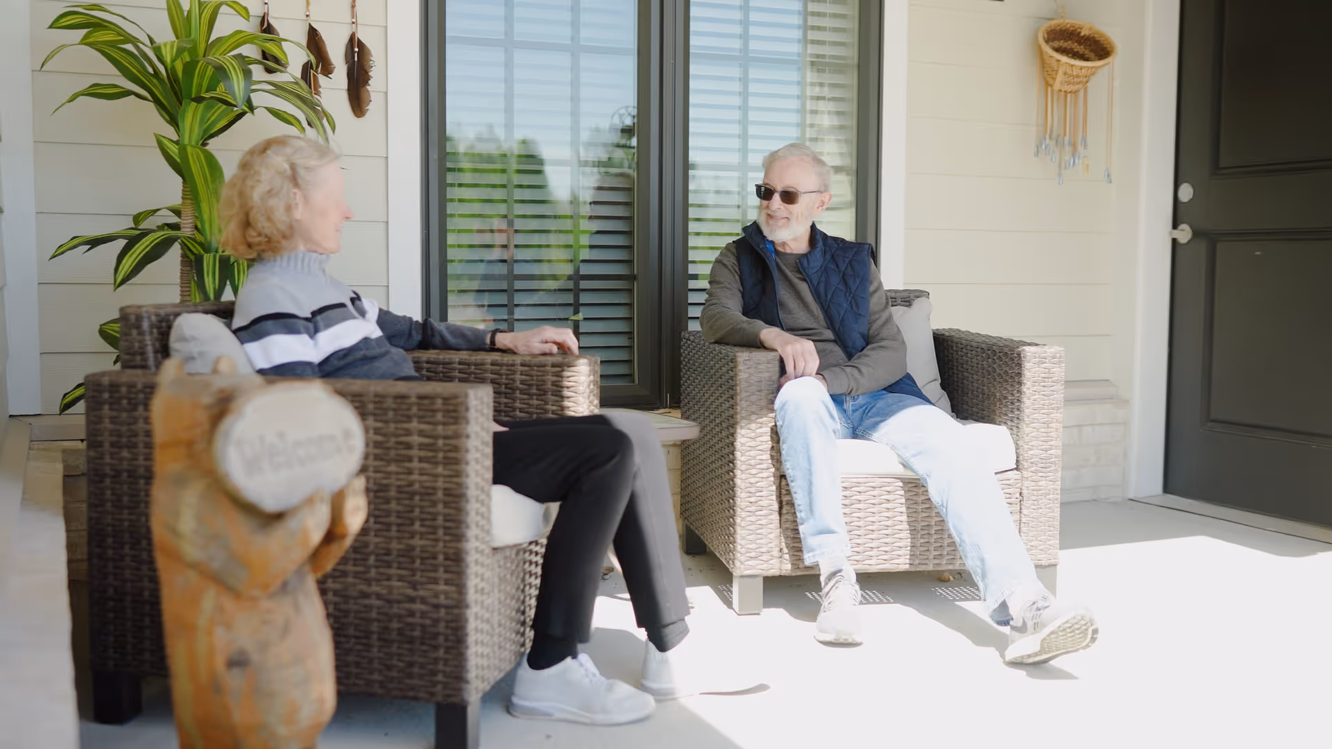 Two elderly people sitting and conversing on a porch with wicker chairs, a potted plant, and a wooden bear statue holding a welcome sign.