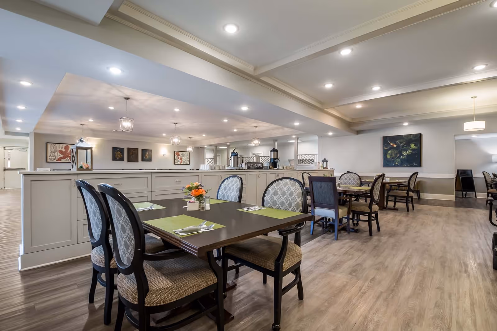 Bright dining room with multiple set tables and upholstered chairs in a senior living facility.