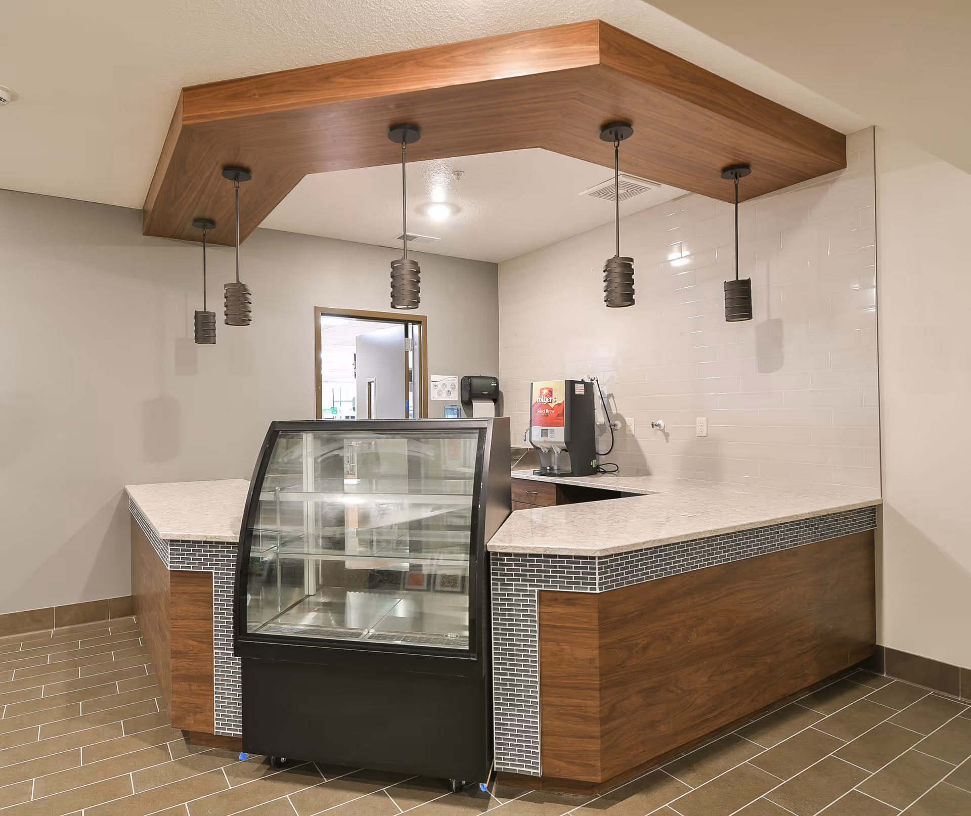 Empty snack bar/cafe counter with a glass display case, pendant lights, and a beverage machine behind the marble-topped serving counter.