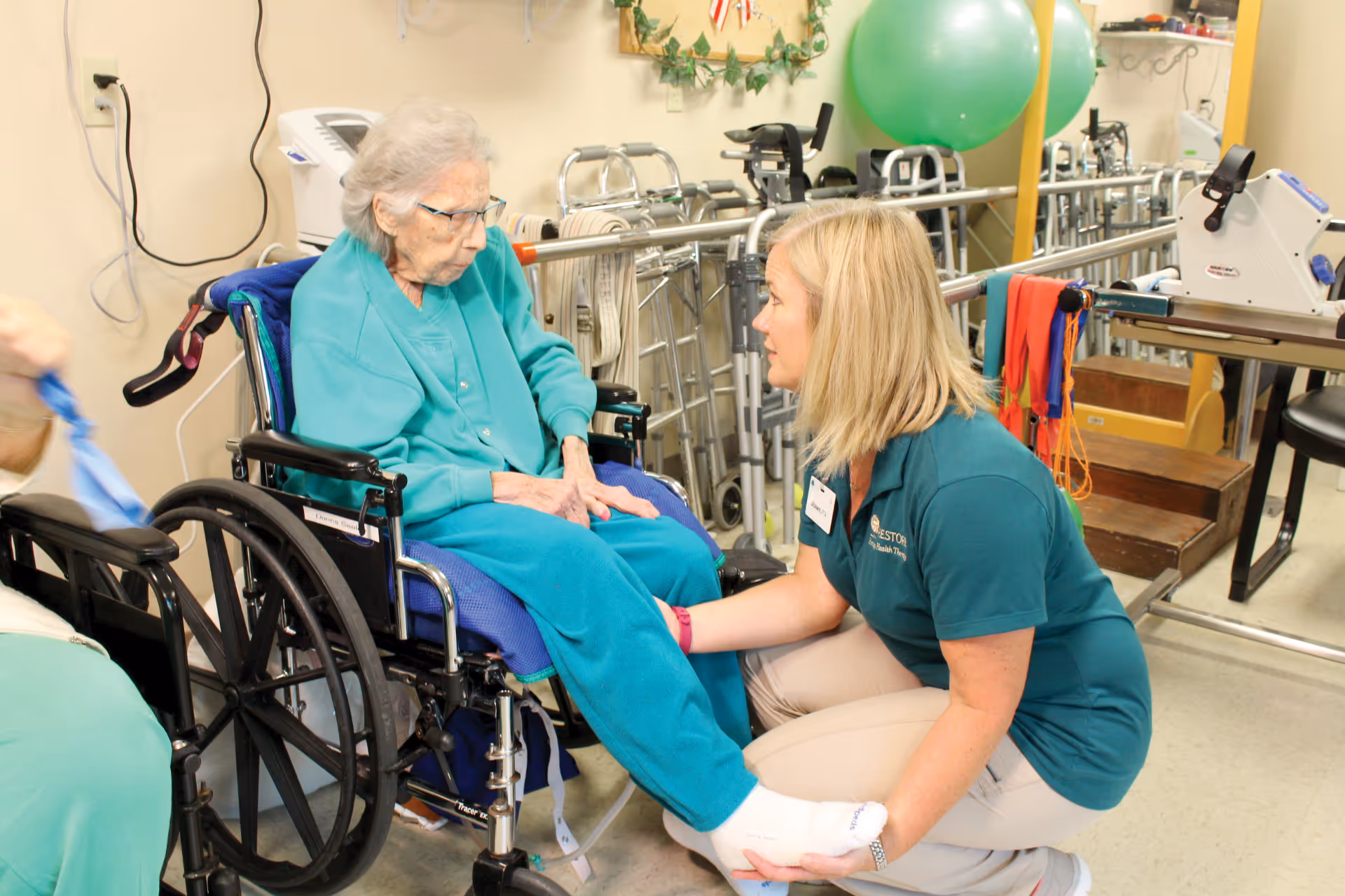 An elderly woman in a wheelchair wearing a teal outfit is being assisted by a female caregiver in a teal polo shirt and beige pants. They are in a rehabilitation or physical therapy room with exercise equipment, walkers, and therapy bands visible in the background.