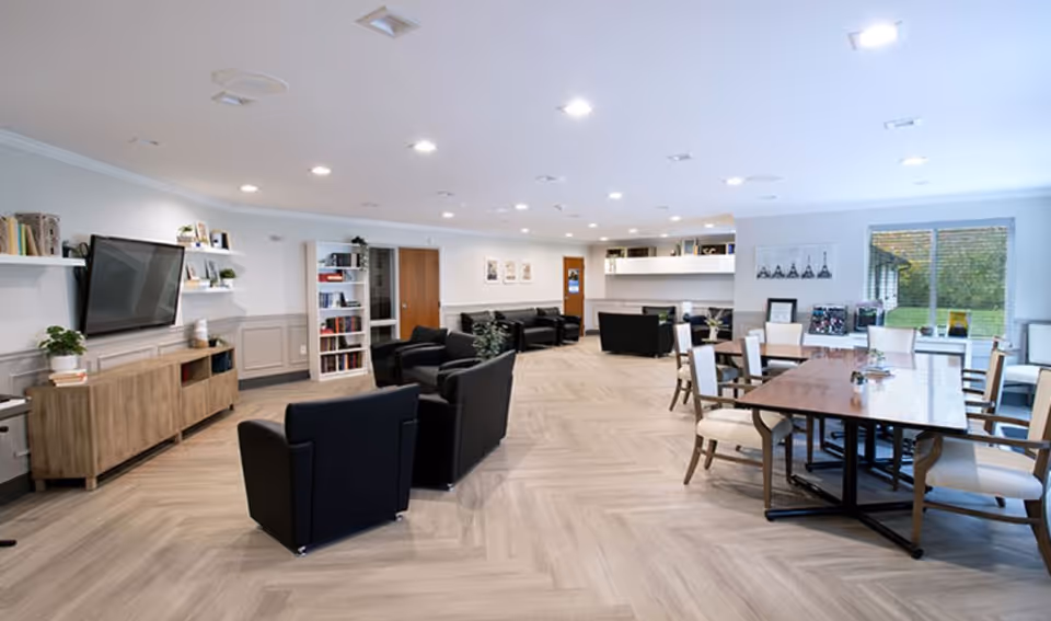Spacious, well-lit communal living area with multiple seating areas, a TV, bookshelves, and a dining table near large windows.