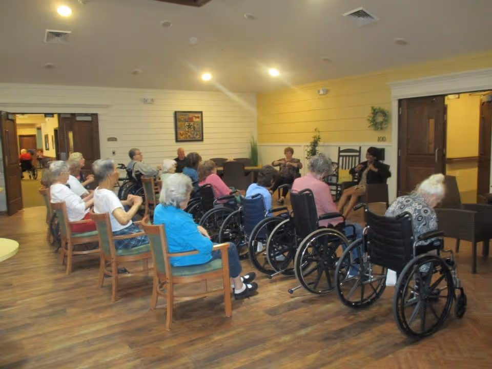 A group of elderly individuals, some in wheelchairs and others seated in chairs, gathered in a spacious, well-lit room with wooden flooring and light-colored walls. Two women appear to be leading an activity or discussion at the front of the group. The room has a welcoming atmosphere with framed artwork on the wall and some plants in the background.