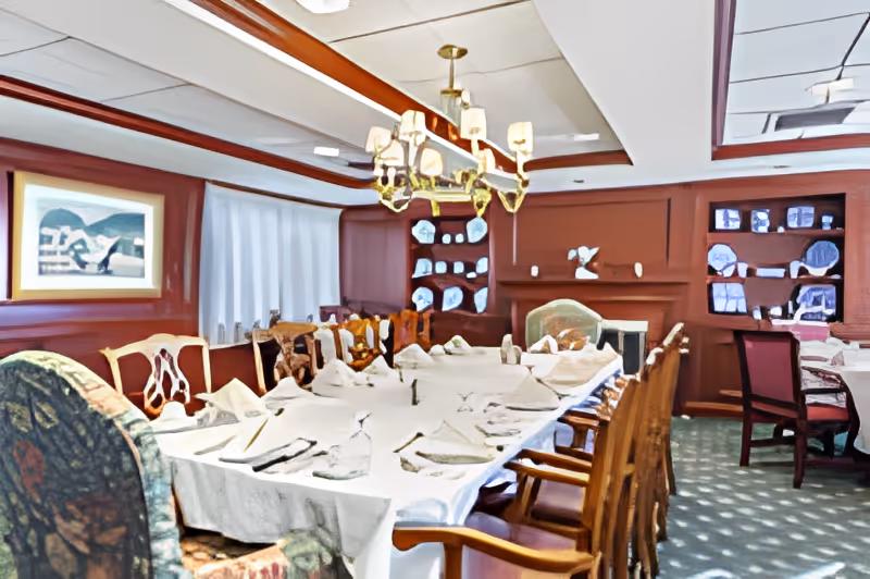 A formal dining room with a long table set with white tablecloth, napkins, glassware, and silverware. The room features wooden chairs, a chandelier hanging from the ceiling, built-in shelves with decorative plates, and a framed painting on the wall.