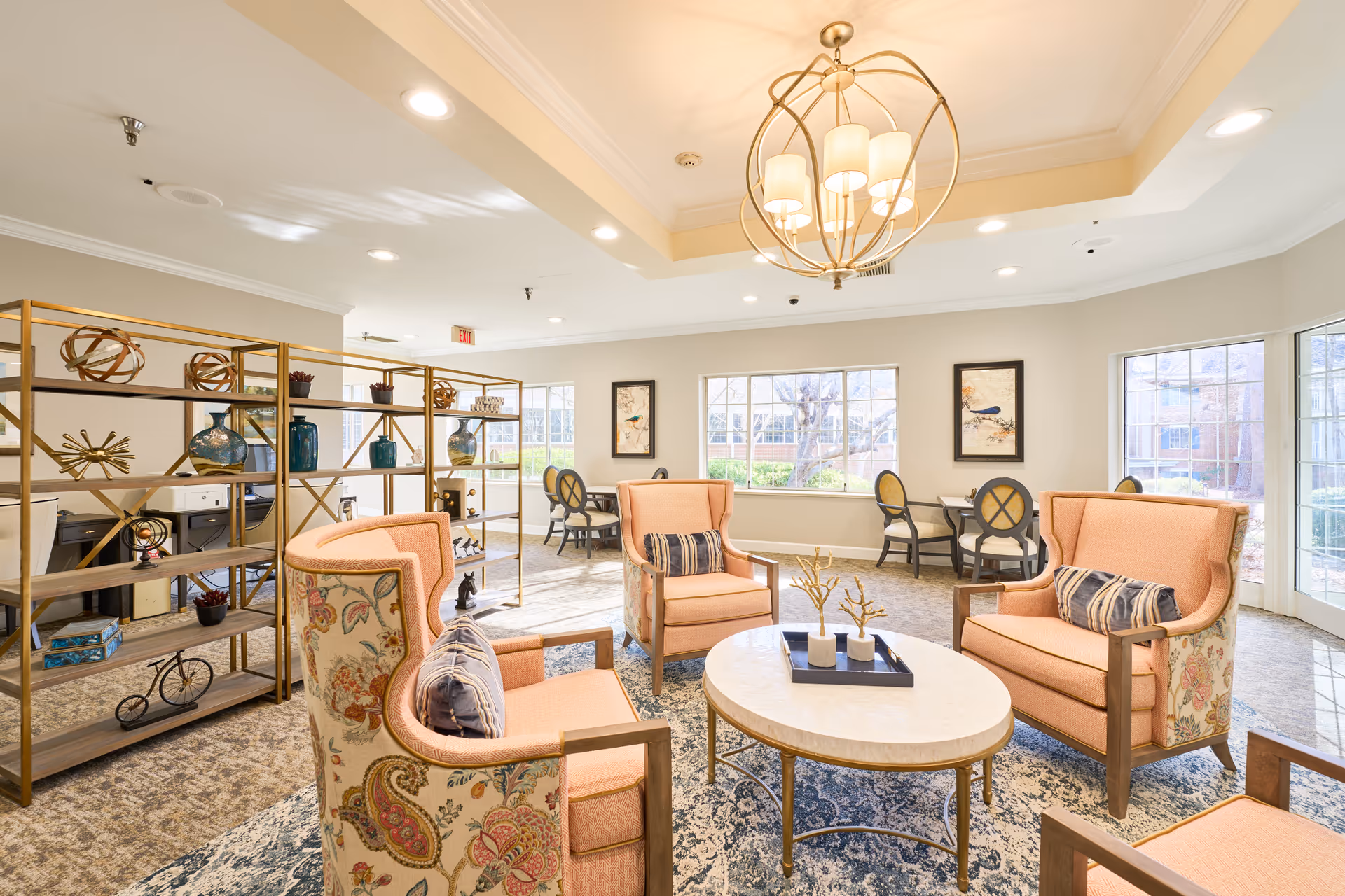 A bright and spacious living room area with four upholstered armchairs arranged around a round coffee table. The chairs have a floral pattern on the back and peach-colored cushions. Behind the seating area is a decorative open shelving unit with various vases and ornaments. Large windows allow natural light to fill the room, and there are small tables with chairs near the windows. A modern chandelier hangs from the ceiling.