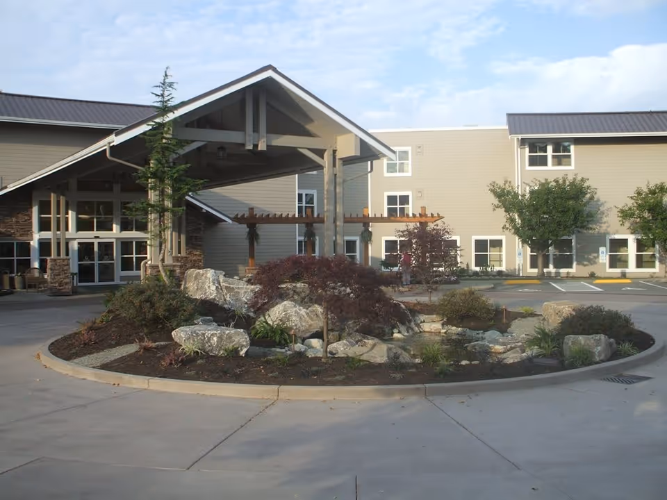 Exterior view of Crista Shores facility showing the main entrance with a covered drop-off area, landscaped roundabout with rocks, plants, and small trees, and a beige multi-story building with several windows in the background.