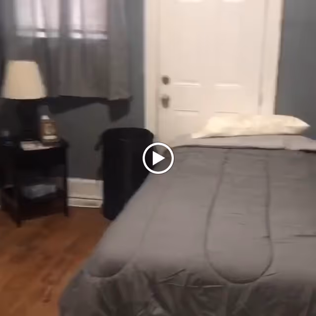 A small bedroom with a single bed covered in a gray comforter and a white pillow. Next to the bed is a black nightstand with a lamp and some items on it. A window with gray curtains is visible on the wall behind the nightstand. The floor is wooden, and there is a white door at the head of the bed.