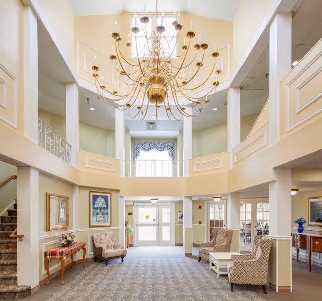 A spacious and elegant lobby area with high ceilings and a large, ornate gold chandelier hanging in the center. The room features beige walls with white trim, carpeted floors, and comfortable seating including armchairs and a small table. There is a staircase on the left side, framed artwork on the walls, and large windows allowing natural light to fill the space.