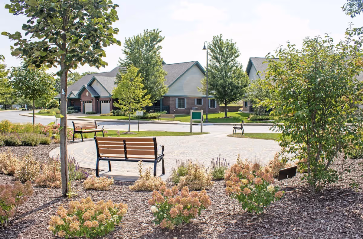 Outdoor garden area with benches, trees, and flowering plants in front of residential buildings with garages under a clear sky.
