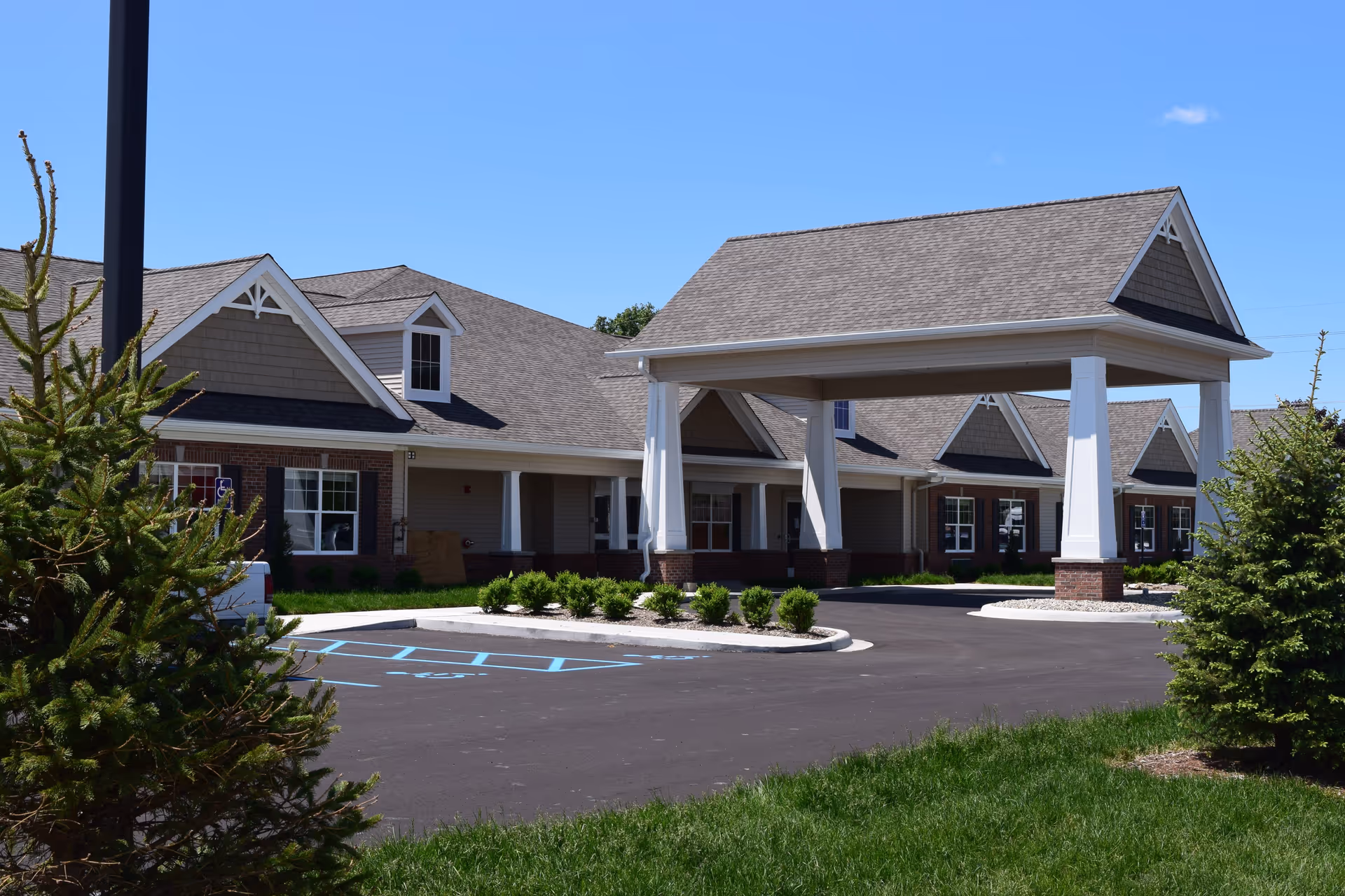 Exterior view of a single-story assisted living facility with a covered entrance, multiple windows, and a parking area with handicapped spaces. The building has a pitched roof with dormer windows and is surrounded by green grass and small trees under a clear blue sky.