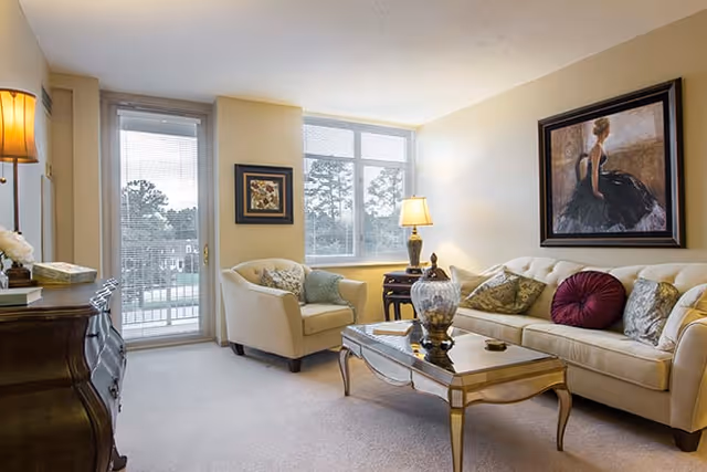 A well-lit living room with a beige sofa adorned with decorative pillows, a matching armchair, a glass-top coffee table with a decorative vase, a side table with a lamp, framed artwork on the walls, and a large window letting in natural light.