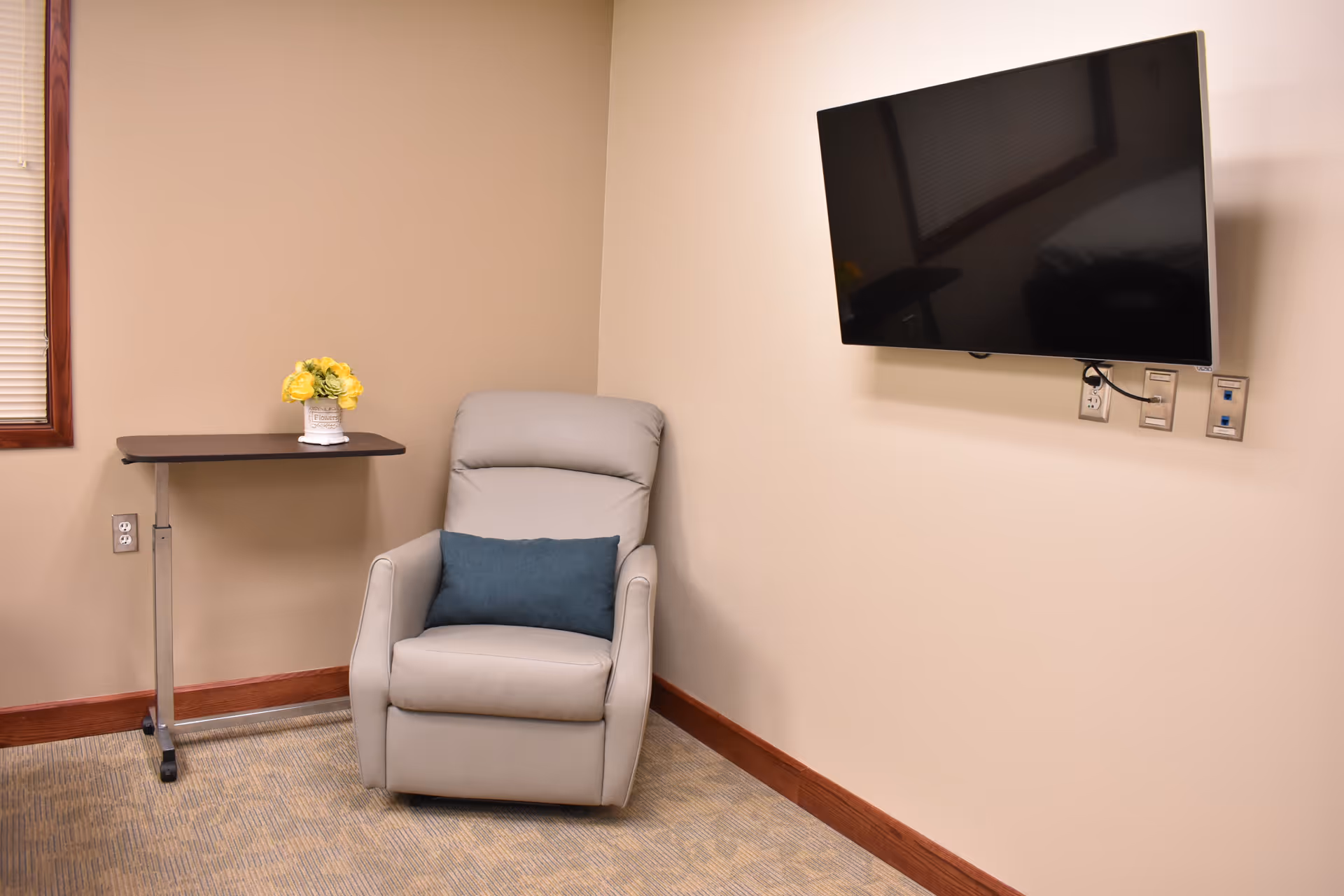 A cozy corner of a room featuring a light gray recliner chair with a blue cushion, a small adjustable table with a vase of yellow flowers, a wall-mounted flat screen TV, beige walls, and a window with closed blinds.