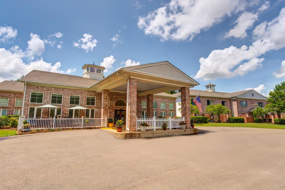 Front exterior view of a senior living facility named Castlewoods Place, featuring a brick building with large windows, a covered entrance with columns, potted plants, and an American flag. The sky is partly cloudy with blue patches.