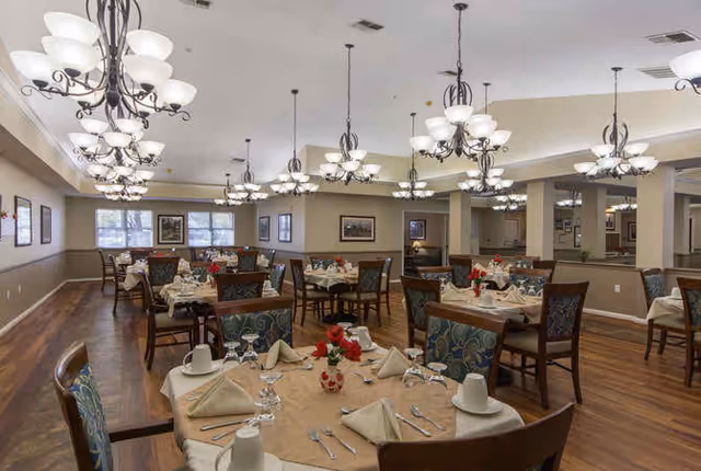 A spacious dining room with multiple round tables set for a meal. Each table is covered with a beige tablecloth and neatly arranged with folded napkins, cups, glasses, and silverware. The chairs have patterned upholstery, and the room is well-lit with multiple elegant chandeliers hanging from the ceiling. The walls are decorated with framed pictures, and large windows allow natural light to enter.
