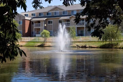 Exterior view of a senior living building beside a pond with a fountain.