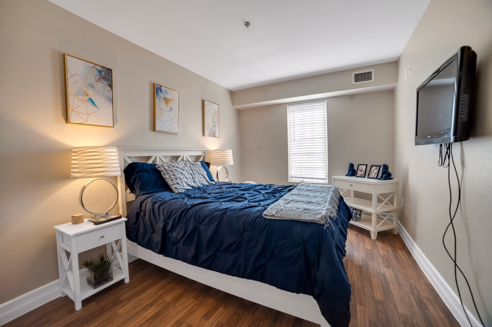 A neatly arranged bedroom with a white bed frame and navy blue bedding. There are two white nightstands on either side of the bed, each with a lamp. Three abstract art pieces hang on the beige wall above the bed. A window with white blinds is on the far wall, and a flat-screen TV is mounted on the right wall. The floor is wooden.