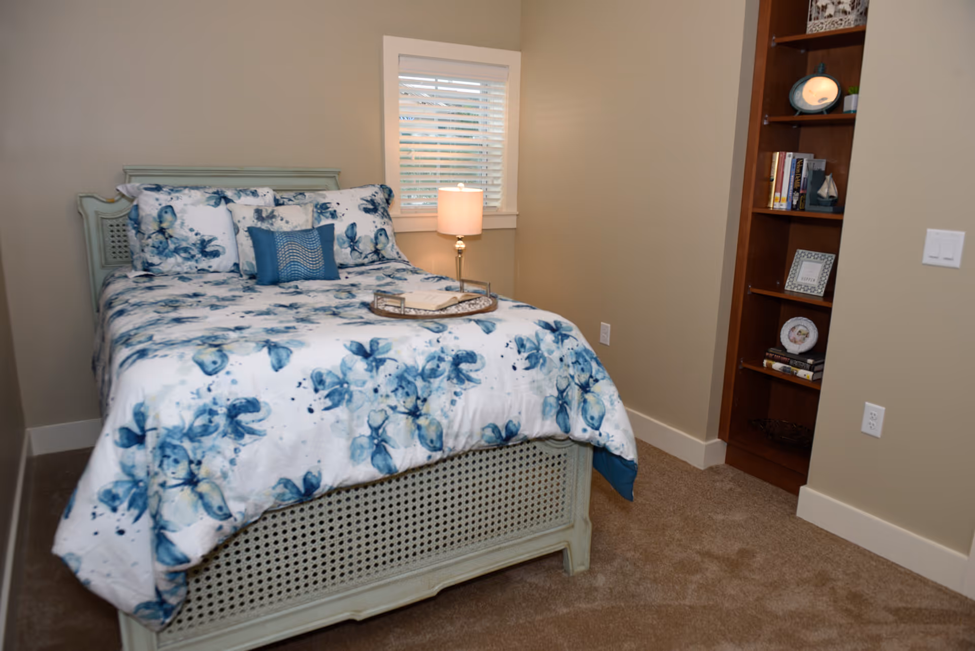 A bedroom with a blue-and-white floral bed, a lit bedside lamp by a small window, and built-in wooden shelving.