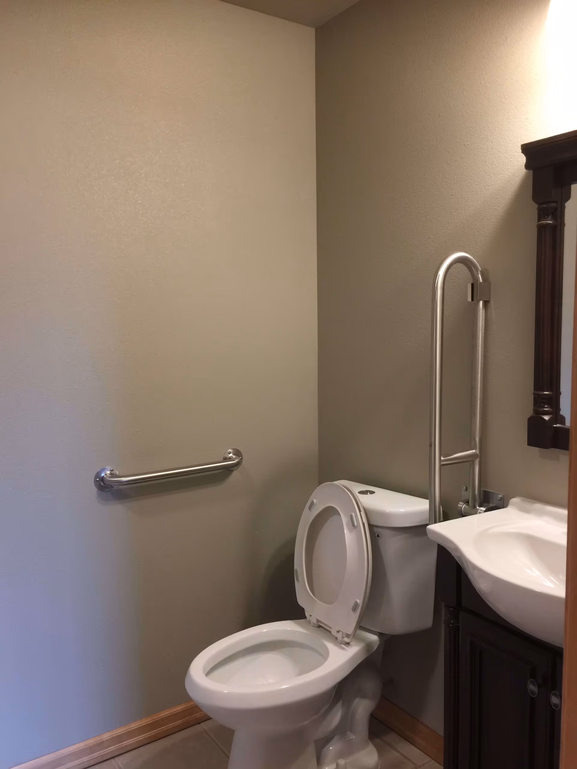 A small bathroom with a white toilet with the seat up, a white sink with a dark wooden cabinet below it, a wall-mounted metal grab bar next to the toilet, and a mirror with a dark wooden frame above the sink.
