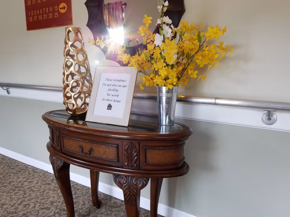 A decorative wooden console table with a glass top against a wall in a hallway. On the table are a metallic vase with yellow and white flowers, a gold abstract sculpture, and a framed sign that reads 'These residents Do not live in our facility. We work In their home.' A mirror with a dark frame hangs above the table, and a handrail runs along the wall behind it.