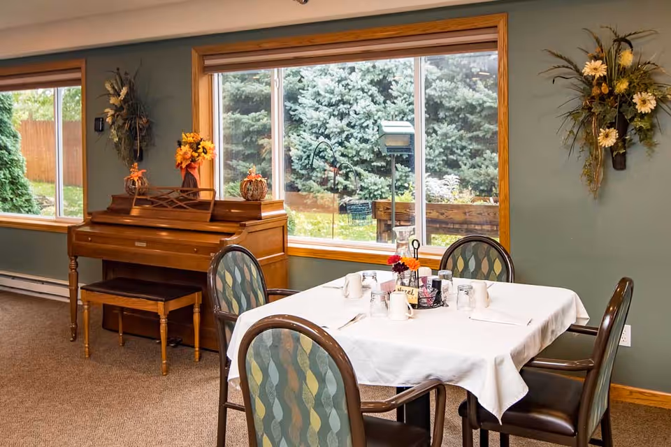 A dining area with a table set for meals and a piano nearby, surrounded by large windows overlooking greenery.