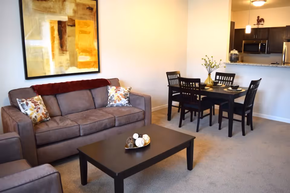 A cozy living and dining area featuring a brown sofa with decorative pillows, a dark wooden coffee table with decorative balls, a dining table set with four chairs and place settings, and a kitchen area with dark cabinets and stainless steel appliances in the background.