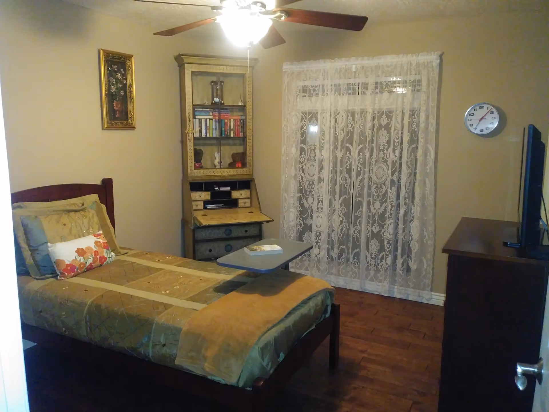 Single-occupancy bedroom with a bed, overbed table, wooden furniture, lace-covered window, and a wall clock.