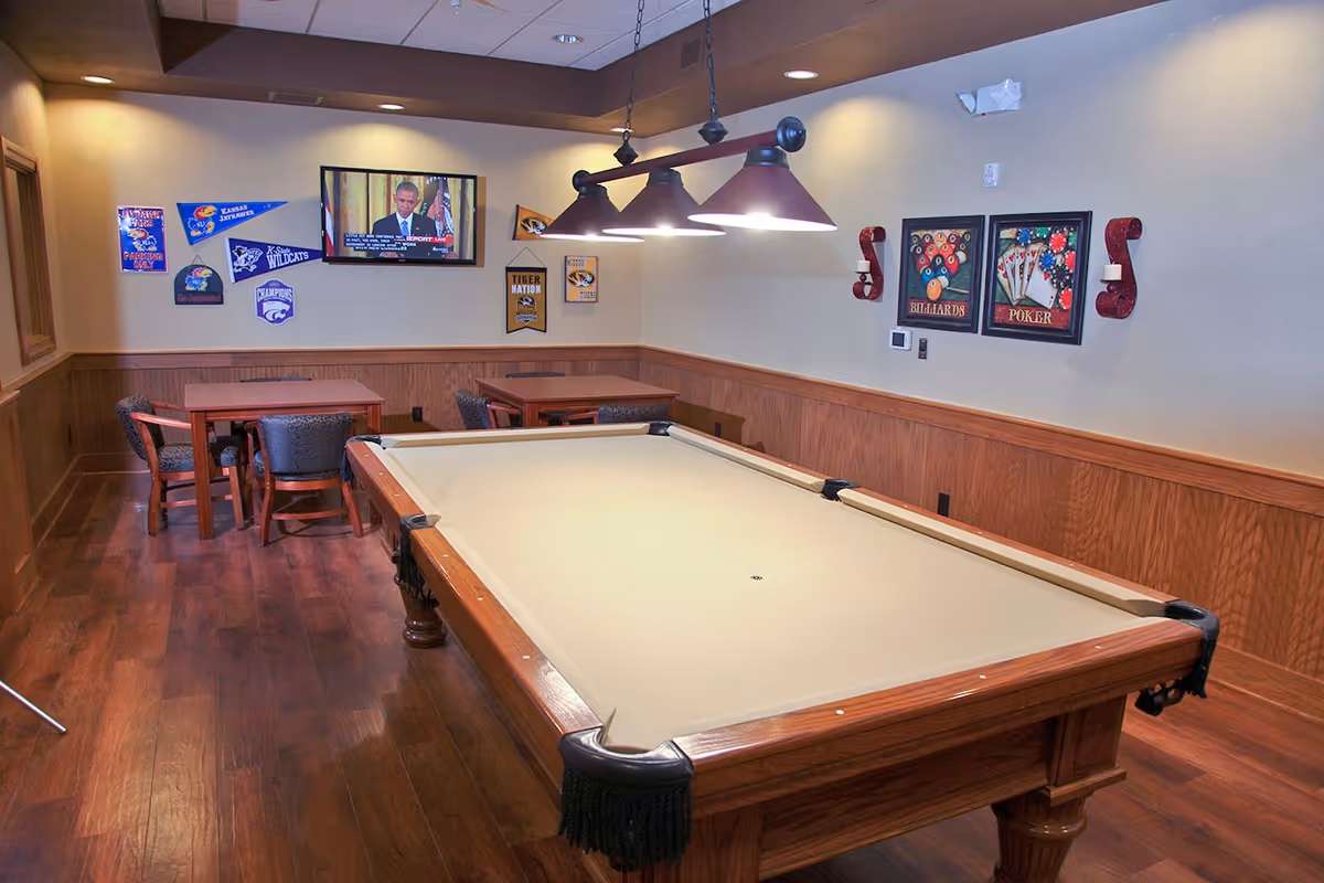 Billiards table in a wood-paneled recreation room with hanging lights, card tables, chairs, wall decor, and a mounted TV.