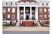 Front exterior view of a large, multi-story brick building with white columns and multiple windows, featuring stairs leading up to the main entrance and a black metal fence in front.
