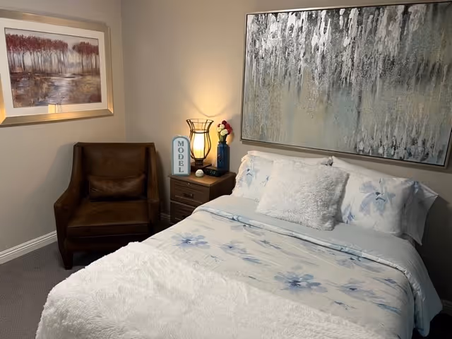A cozy bedroom featuring a bed with white and blue floral bedding, a fluffy white pillow, and a large abstract painting above the headboard. To the left of the bed is a wooden nightstand with a lamp, a blue vase with flowers, and a decorative sign that reads 'MODEL'. A brown armchair is positioned in the corner under a framed landscape painting.