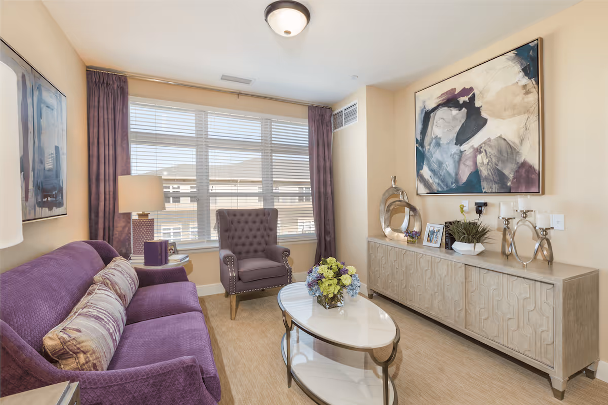 A cozy living room with a purple sofa and matching armchair, a white oval coffee table with a flower arrangement, a sideboard with decorative items including a modern sculpture, framed photos, and candle holders, and two abstract paintings on the walls. The room has beige walls, a large window with blinds and purple curtains, and a ceiling light fixture.