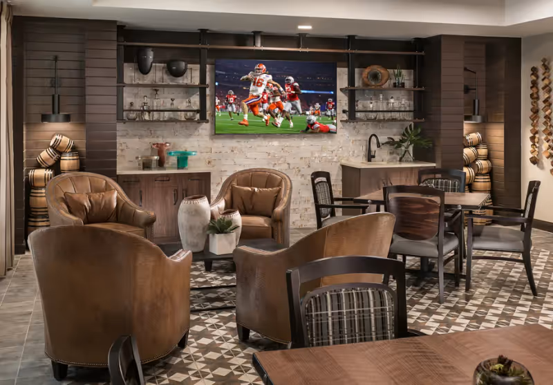 A cozy common area with four brown leather armchairs arranged around a small table with decorative vases and a plant. Behind the chairs is a wall with shelves holding glassware and decorative items, and a flat-screen TV mounted on a white brick wall showing a football game. To the right, there are tables and chairs for dining or socializing, with a small sink and countertop area. The room features warm lighting and patterned tile flooring.