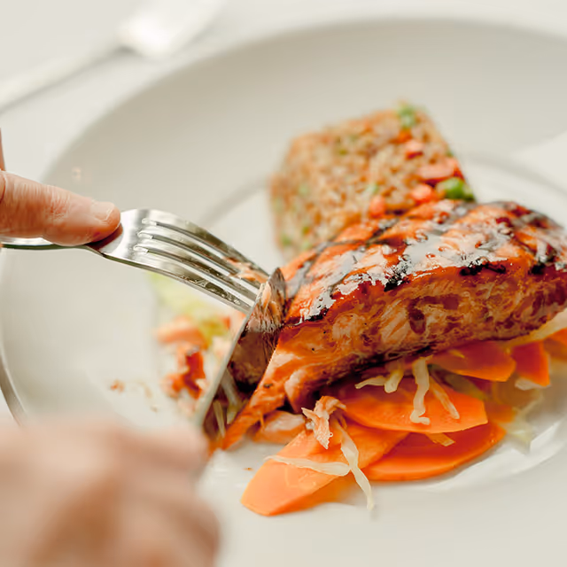 Close-up of a person cutting into a grilled piece of salmon served on a bed of sliced carrots and cabbage, with a side of mixed rice on a white plate.
