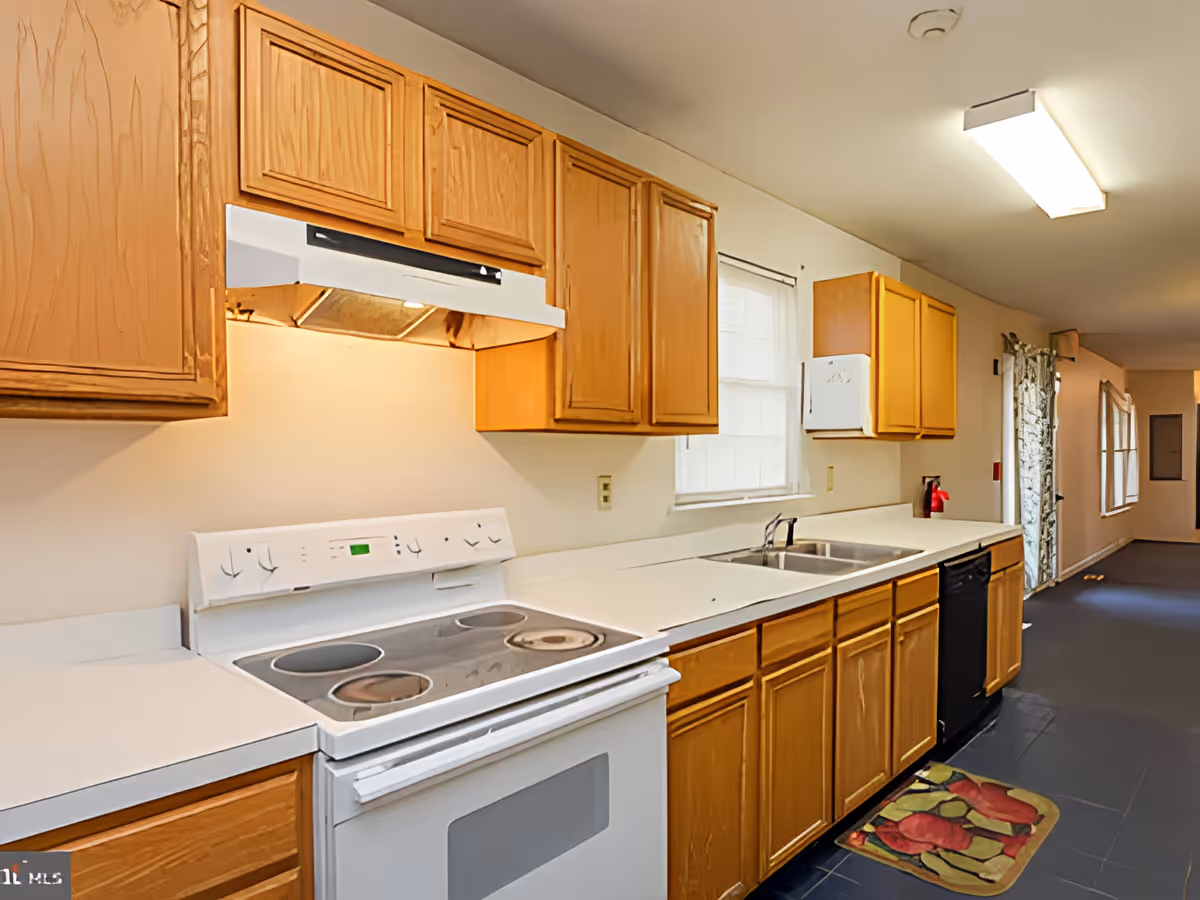 Long galley-style kitchen with oak cabinets, a white electric stove, double sink under a window, and dark tile flooring.