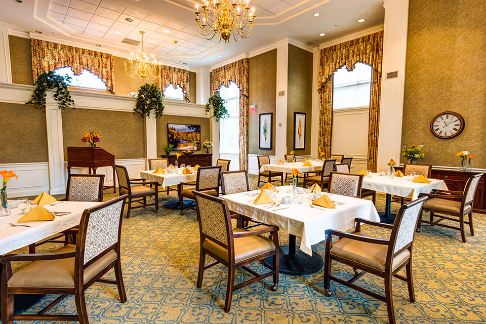 Well-lit dining room with tables set with white tablecloths and yellow napkins, wooden chairs, chandeliers, and decorative curtains.