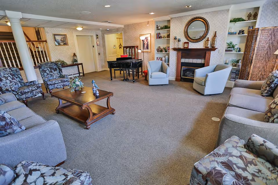 A spacious living room area with multiple upholstered chairs and sofas arranged around a wooden coffee table. There is a fireplace with a round mirror above it, built-in shelves with decorative items, a black grand piano, and a staircase with white railings in the background.