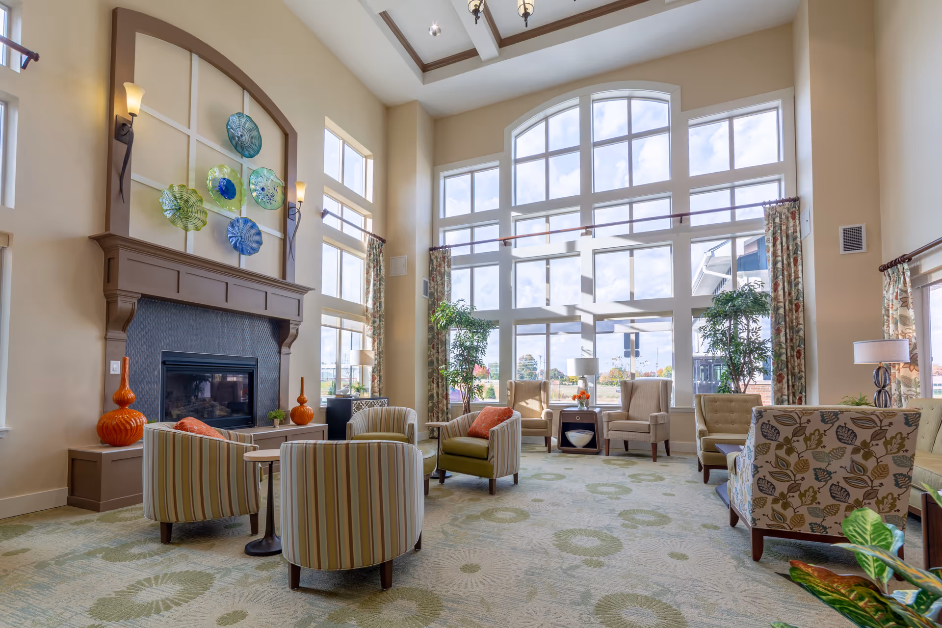 A spacious and bright living room area with large floor-to-ceiling windows letting in natural light. The room features a fireplace with decorative glass art above it, several upholstered chairs with striped and floral patterns arranged around small tables, potted plants, and tall curtains framing the windows.