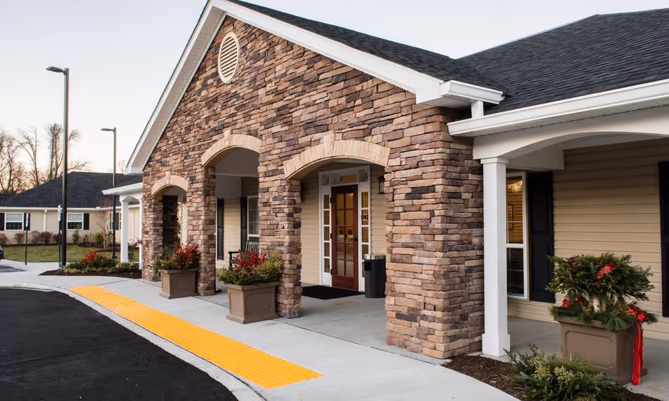 Exterior view of a senior living facility entrance with stone pillars, a covered porch, potted plants with red flowers, and a sidewalk with a yellow tactile paving strip.
