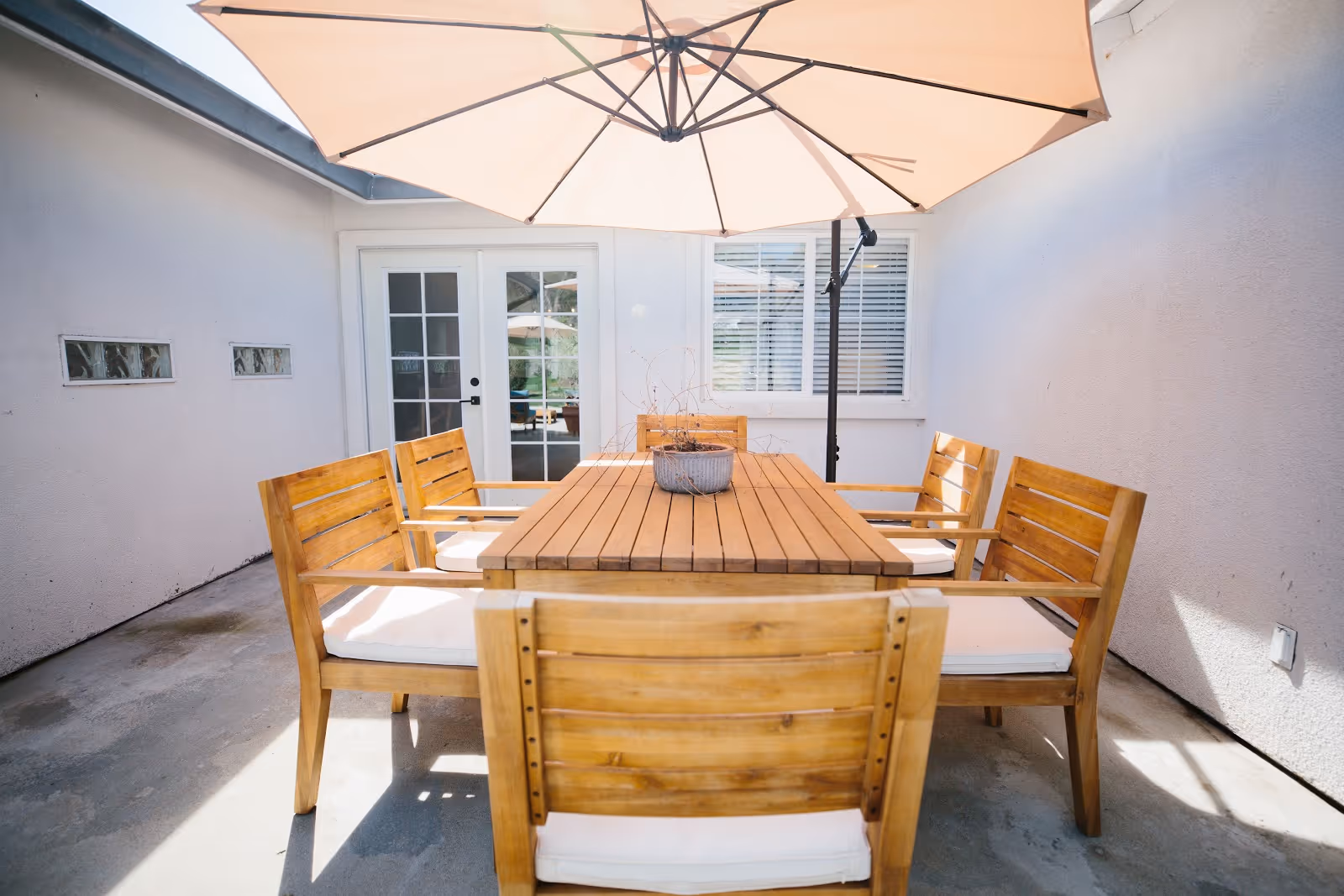 A wooden outdoor dining table surrounded by six cushioned chairs under a large patio umbrella in a sunlit courtyard in front of glass doors.