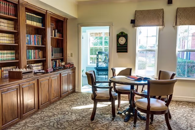 A cozy room with a round wooden table surrounded by four cushioned chairs. To the left, there is a large wooden bookshelf filled with books and decorative items. The room has carpeted flooring with a patterned design. Two large windows on the right wall let in natural light, and a wall clock is mounted between the windows. Through an open doorway, another room with a chair and large windows is visible.