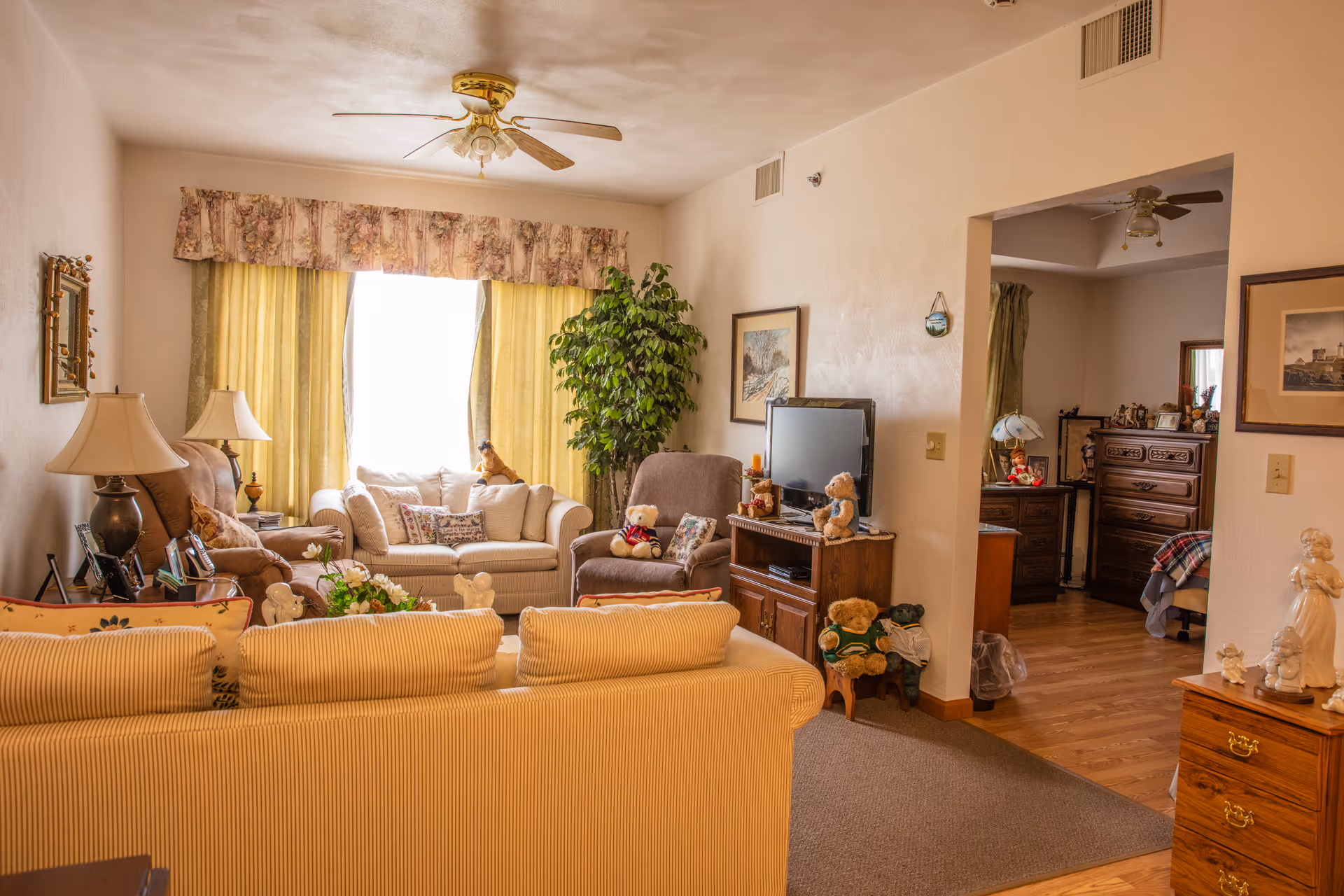 A cozy living room with beige walls and a ceiling fan. The room features a beige striped sofa in the foreground, a brown recliner, and a cream-colored loveseat near a window with yellow curtains and a floral valance. There are several teddy bears placed on the furniture and a wooden TV stand with a flat-screen TV. A large green potted plant is next to the loveseat. The room opens into a bedroom area with wooden furniture and a bed visible.