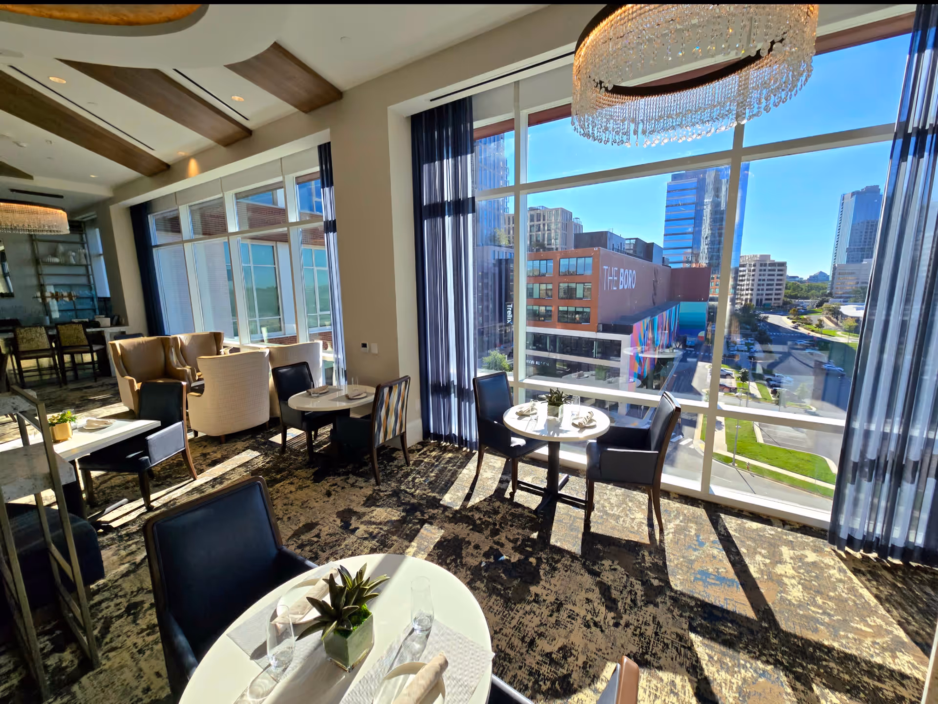 A bright dining room with large floor-to-ceiling windows offering a view of a cityscape. The room features several round tables set with plates, glasses, and napkins, surrounded by a mix of black and beige chairs. A large chandelier hangs from the ceiling, and sheer curtains frame the windows.